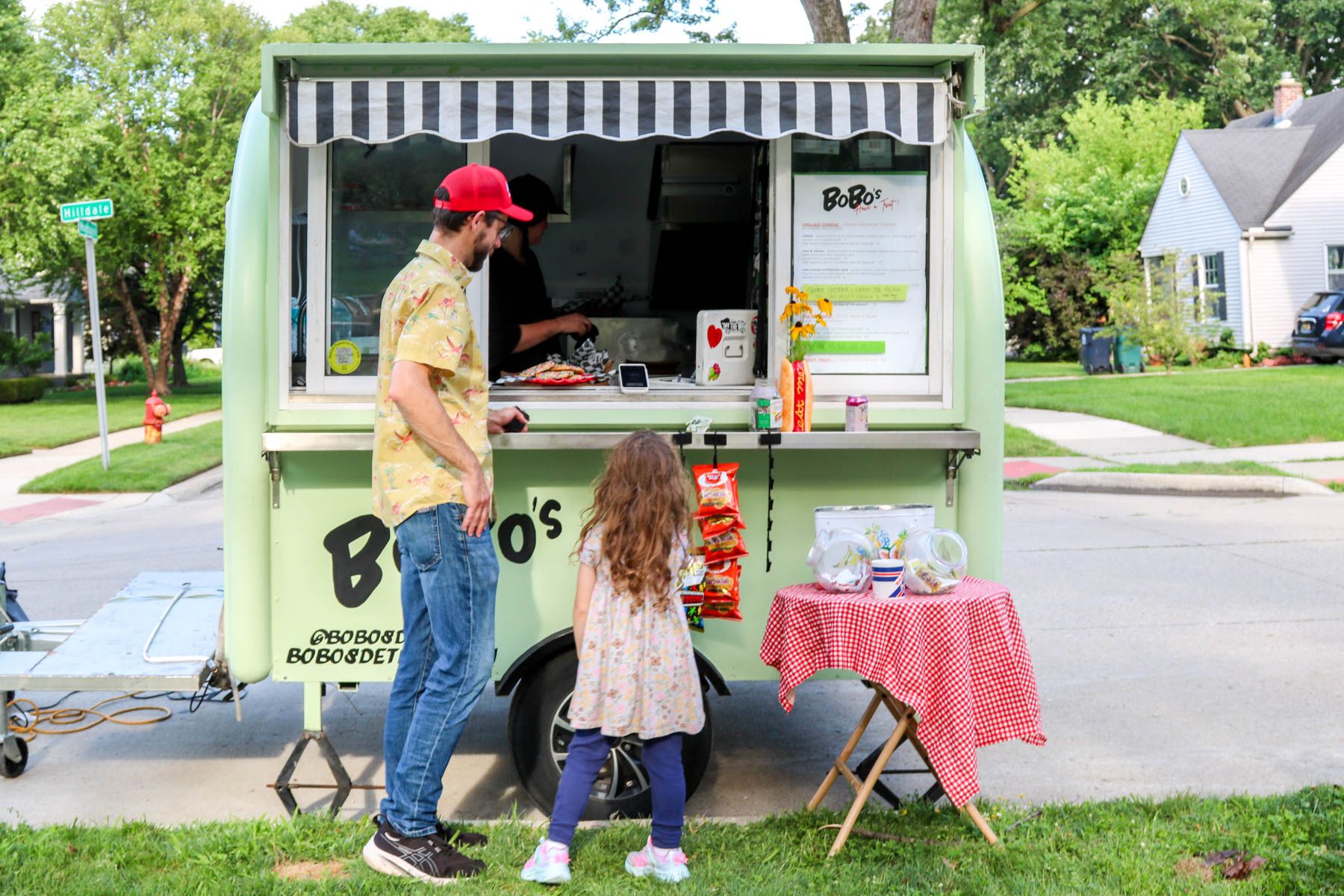 A dad and his daughter ordering at a mint-green food truck on a tree-lined street