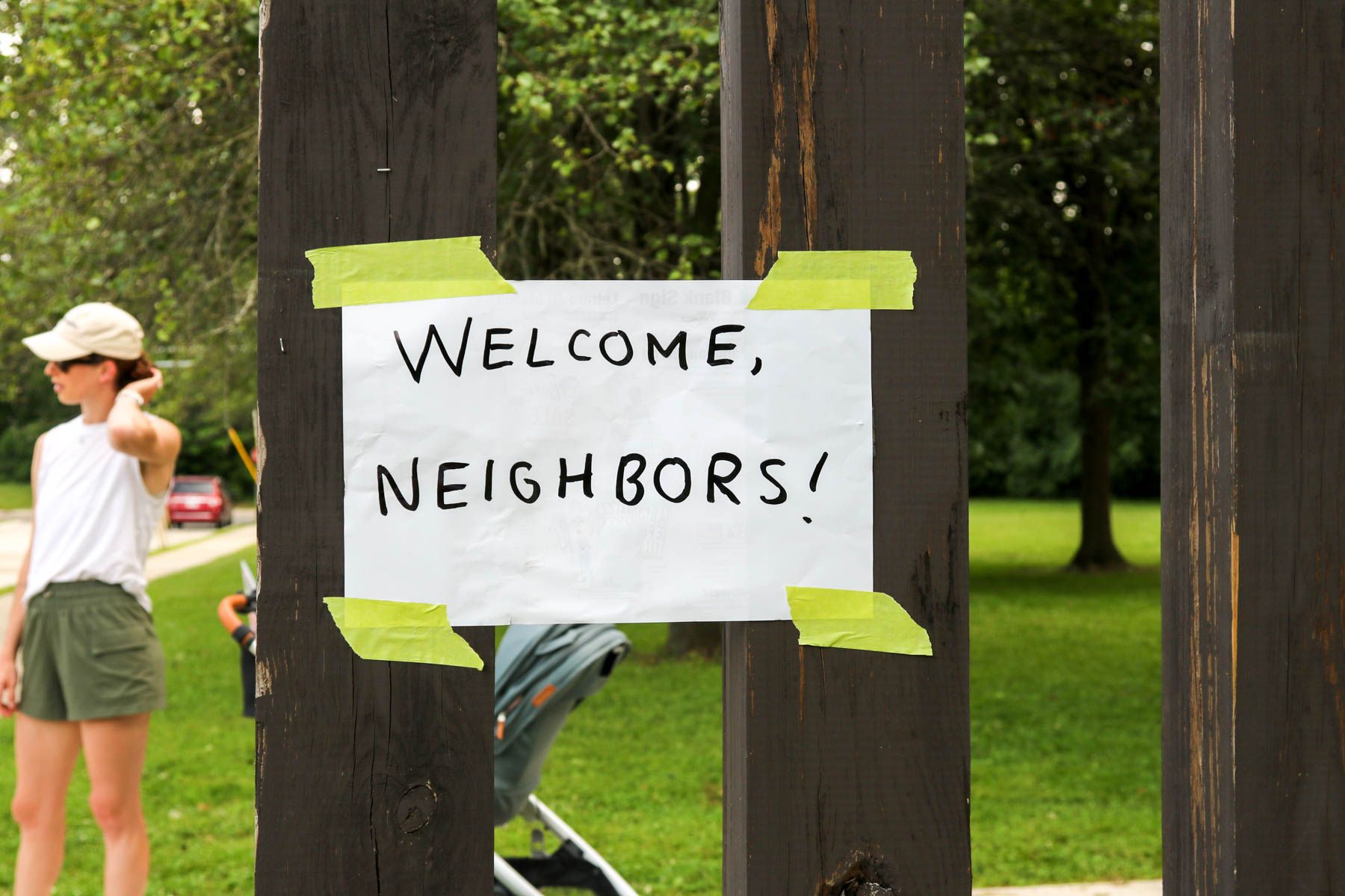 A hand-lettered “Welcome, Neighbors!” sign taped to a wooden post in the park